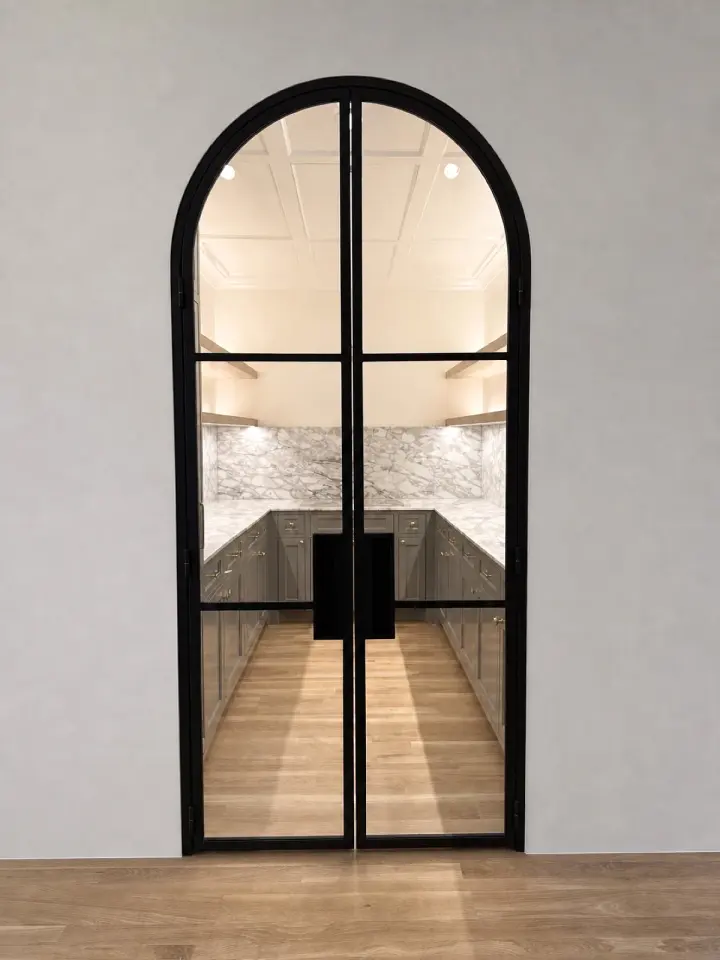 The finished full-arch black steel pantry pair seen from the kitchen, marble counters beyond the glass