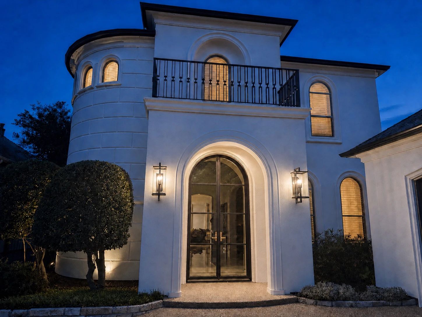 Arched iron double entry door lit from within at dusk, with a balcony railing above and a wall lantern to either side